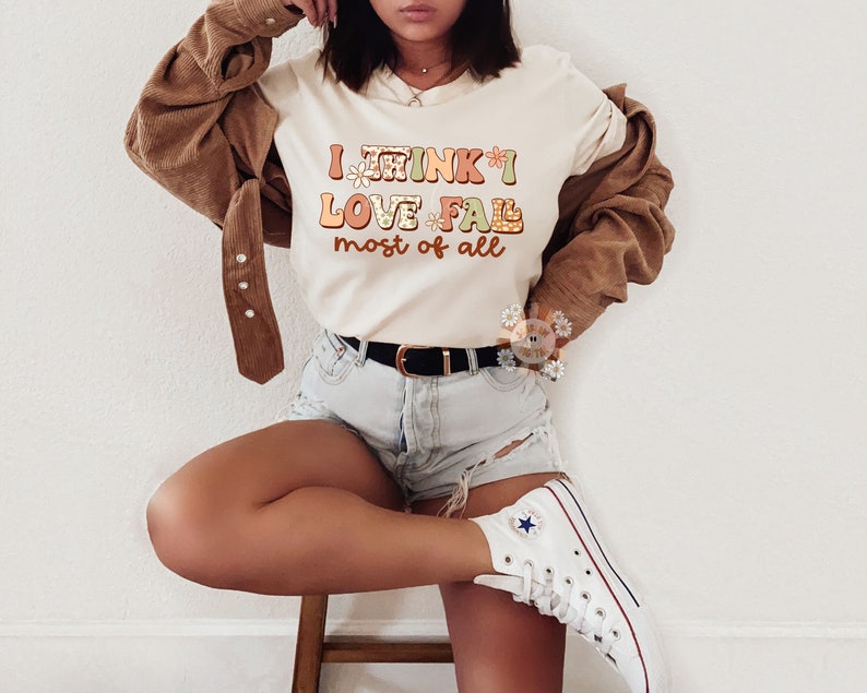 Op de afbeelding: Een wit T-shirt met de tekst "I think I love fall most of all" in kleurrijke, gestileerde letters met bloemmotieven.