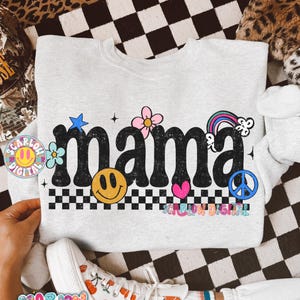 May include: A light gray sweatshirt with the word "mama" in black, decorated with a smiley face, flowers, a rainbow, and a peace sign. The sweatshirt has a checkered pattern at the bottom of the word.