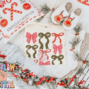 May include: A white t-shirt with a festive design of red and green bows. The shirt is surrounded by holly, a white knitted stocking, and a pillow with the text "Kringle Candy Co."