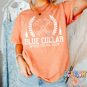 May include: A peach colored t-shirt with a white graphic design of two crossed hammers, a screwdriver, and a wreath. The text "Blue Collar Wives Social Club" is printed below the graphic.