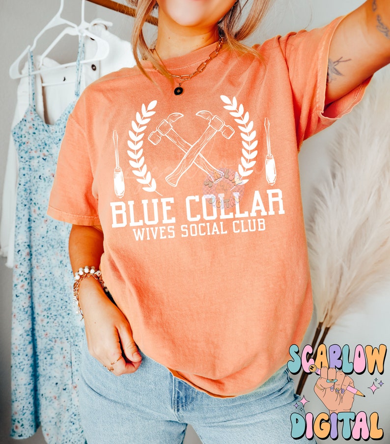 May include: A peach colored t-shirt with a white graphic design of two crossed hammers, a screwdriver, and a wreath. The text "Blue Collar Wives Social Club" is printed below the graphic.