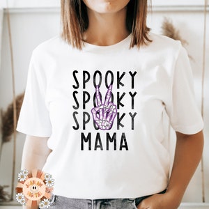 K&ouml;nnte beinhalten: Wei&szlig;es T-Shirt mit schwarzem Text, der "Spooky Spooky Spooky Mama" lautet, mit einer lila Skeletthand, die in der Mitte des Textes ein Peace-Zeichen macht.