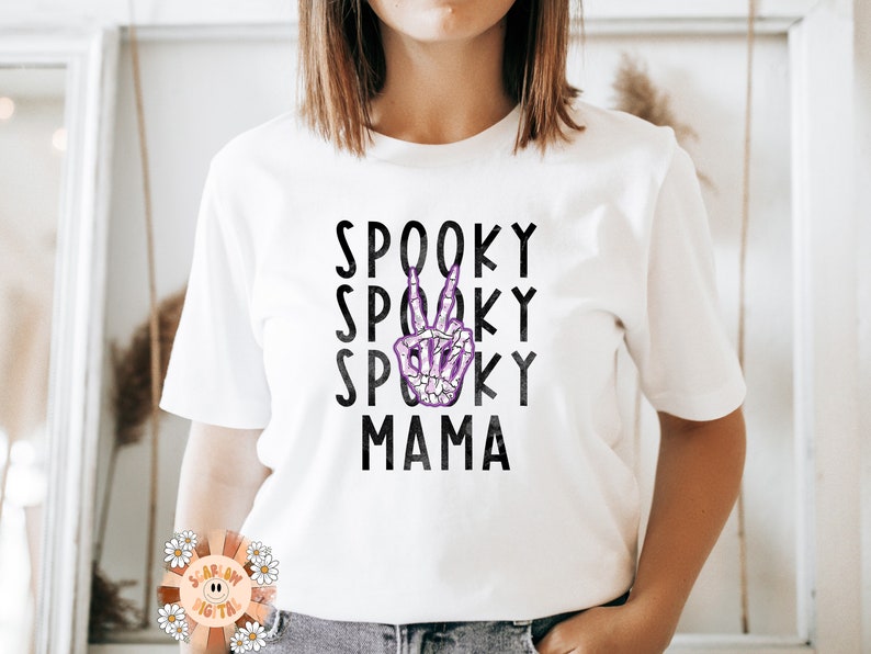 K&ouml;nnte beinhalten: Wei&szlig;es T-Shirt mit schwarzem Text, der "Spooky Spooky Spooky Mama" lautet, mit einer lila Skeletthand, die in der Mitte des Textes ein Peace-Zeichen macht.