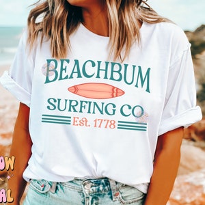 May include: White t-shirt with a teal and pink graphic design that says "Beachbum Surfing Co. Est. 1778".