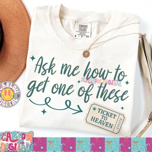 May include: Cream-colored t-shirt with the text "Ask me how to get one of these" in teal script. A "Ticket to Heaven" graphic is on the lower right. A gold necklace and a brown hat are also visible.