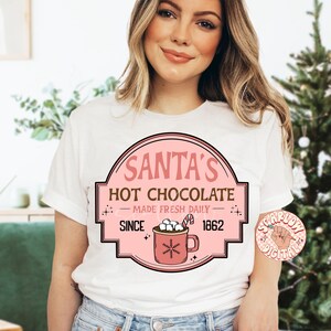 Op de afbeelding: Wit T-shirt met een roze en bruin grafisch ontwerp met de tekst "Santa's Hot Chocolate - Made Fresh Daily - Since 1862". De afbeelding toont een mok met warme chocolademelk met marshmallows.