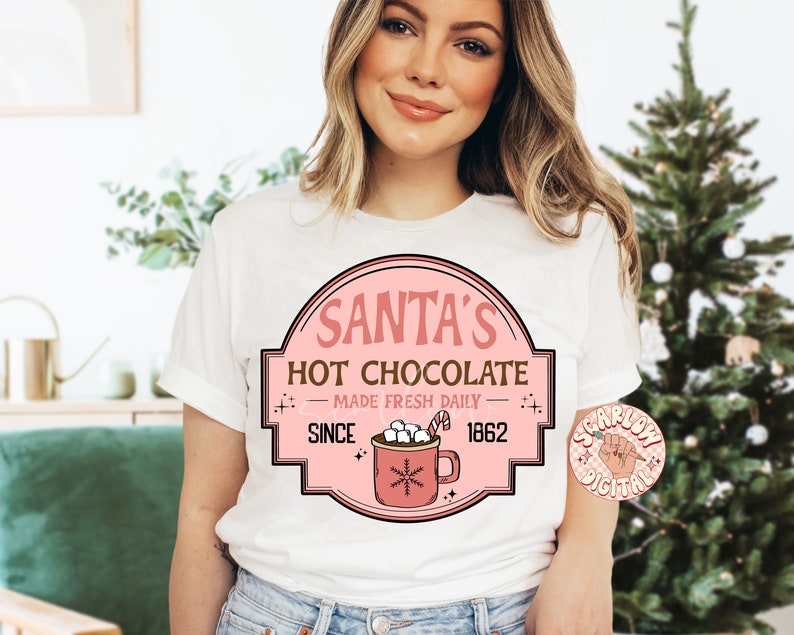 Op de afbeelding: Wit T-shirt met een roze en bruin grafisch ontwerp met de tekst "Santa's Hot Chocolate - Made Fresh Daily - Since 1862". De afbeelding toont een mok met warme chocolademelk met marshmallows.