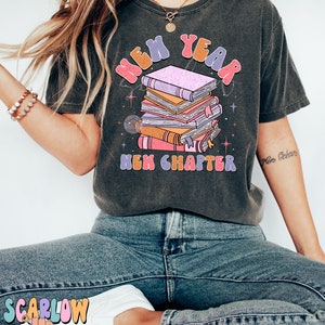 May include: A dark grey t-shirt featuring a graphic of a stack of books and the text "New Year New Chapter" in colourful, retro-style lettering.