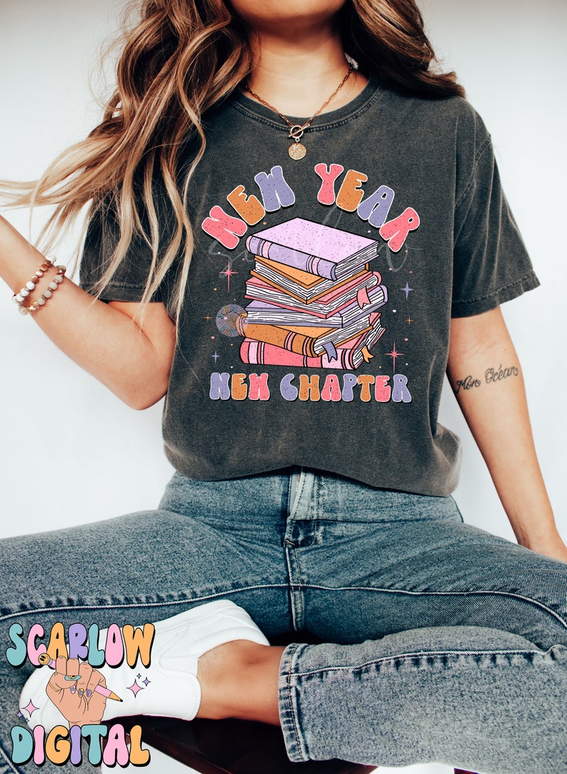 May include: A dark grey t-shirt featuring a graphic of a stack of books and the text "New Year New Chapter" in colourful, retro-style lettering.