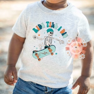 K&ouml;nnte beinhalten: Ein hellgraues T-Shirt mit einem Skelett, das Skateboard f&auml;hrt, und dem Text "Ride Till I Die" in bunten Buchstaben. Das Skelett tr&auml;gt einen blauen Hut und das Skateboard ist blau und orange.