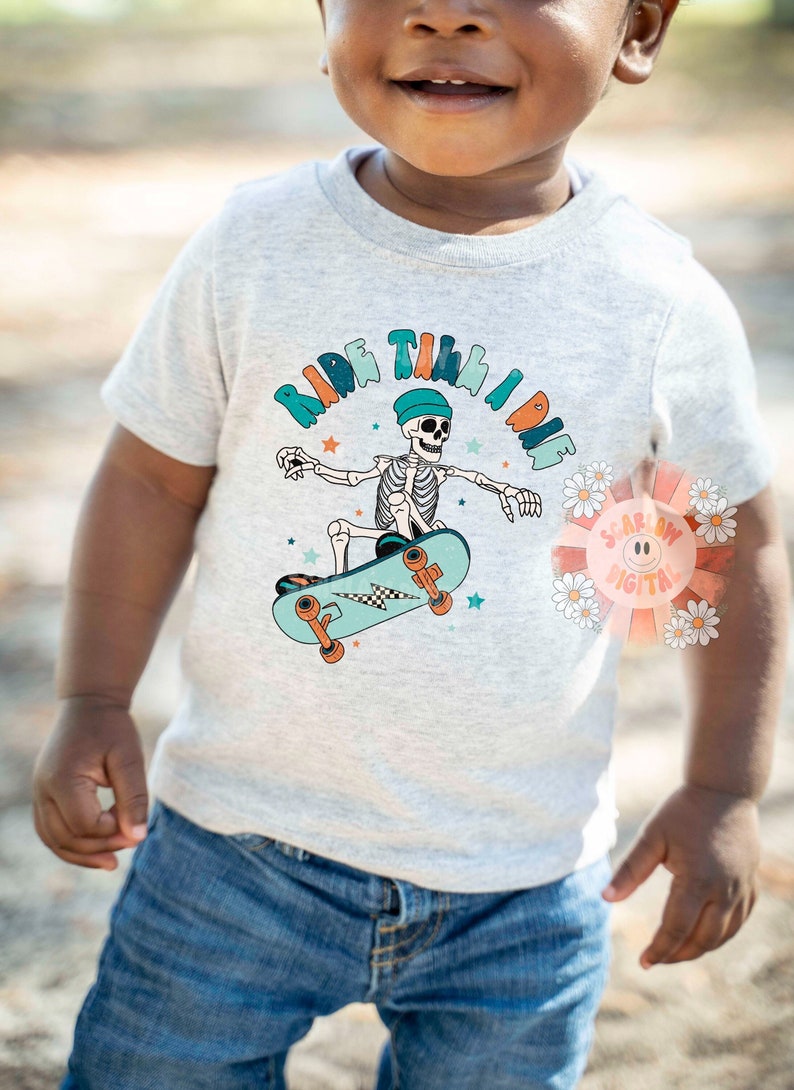 K&ouml;nnte beinhalten: Ein hellgraues T-Shirt mit einem Skelett, das Skateboard f&auml;hrt, und dem Text "Ride Till I Die" in bunten Buchstaben. Das Skelett tr&auml;gt einen blauen Hut und das Skateboard ist blau und orange.