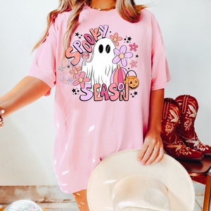 Pu&ograve; includere: Maglietta rosa con un fantasma bianco, fiori e il testo "Spooky Season" in lettere rosa e arancioni.