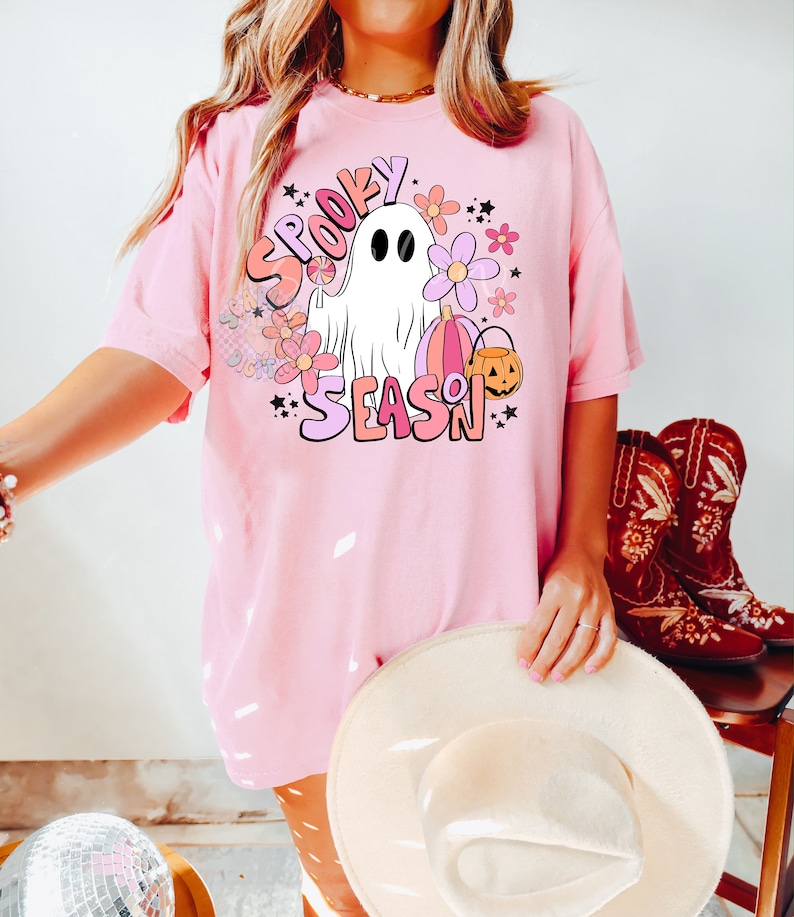 Pu&ograve; includere: Maglietta rosa con un fantasma bianco, fiori e il testo "Spooky Season" in lettere rosa e arancioni.