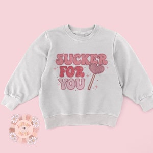 Może przedstawiać: Biała bluza z nadrukiem w kolorze r&oacute;żowym i czerwonym, na kt&oacute;rym widnieje napis "SUCKER FOR YOU" oraz grafika r&oacute;żowego lizak w kształcie serca.