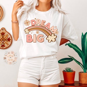 May include: White t-shirt with a rainbow and the text "Dream Big" in pink and brown letters. The rainbow has pink and brown stripes and is decorated with small flowers.