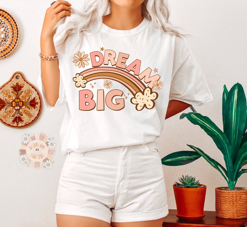 May include: White t-shirt with a rainbow and the text "Dream Big" in pink and brown letters. The rainbow has pink and brown stripes and is decorated with small flowers.