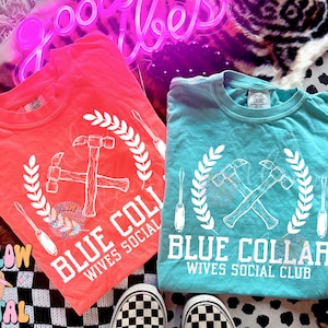 May include: Two t-shirts with a white graphic design of crossed hammers with a wreath and the text "Blue Collar Wives Social Club". One shirt is coral pink and the other is turquoise.