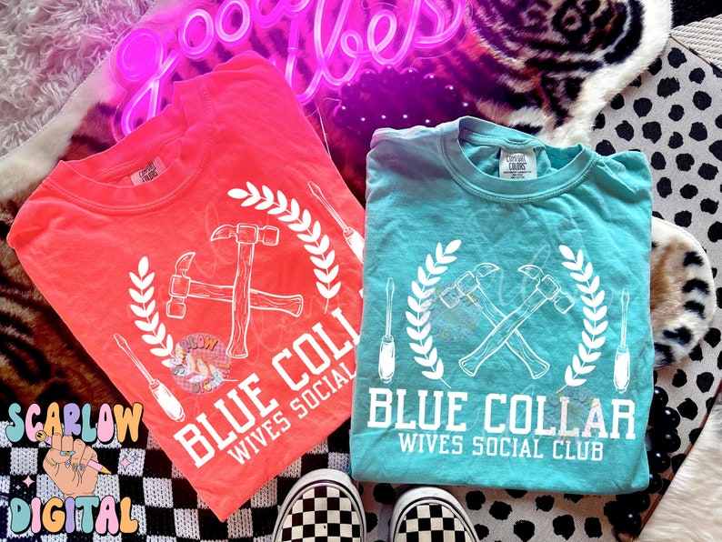May include: Two t-shirts with a white graphic design of crossed hammers with a wreath and the text "Blue Collar Wives Social Club". One shirt is coral pink and the other is turquoise.