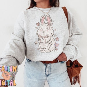 May include: A gray sweatshirt with a cute cartoon bunny wearing a pink bow and surrounded by pink flowers. The sweatshirt is being worn by a person with blue jeans and a brown fringed bag.