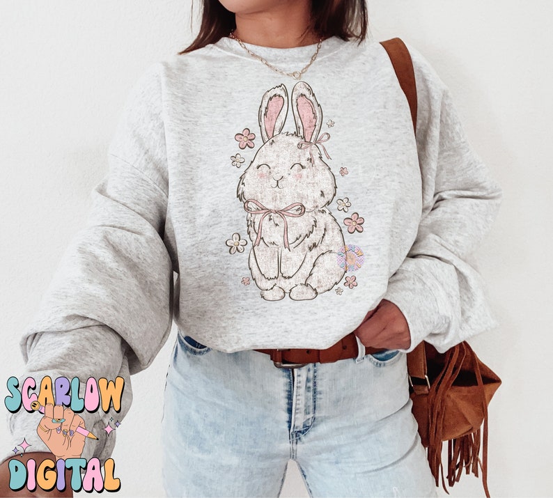 May include: A gray sweatshirt with a cute cartoon bunny wearing a pink bow and surrounded by pink flowers. The sweatshirt is being worn by a person with blue jeans and a brown fringed bag.
