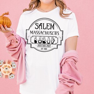 Może przedstawiać: Biała koszulka z czarno-białym nadrukiem graficznym z napisem "Salem Massachusetts Apothecary Est. 1692".