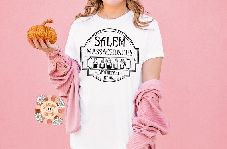 Może przedstawiać: Biała koszulka z czarno-białym nadrukiem graficznym z napisem "Salem Massachusetts Apothecary Est. 1692".