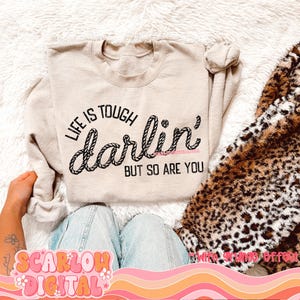May include: A beige sweatshirt with the text "Life is tough darlin' but so are you" printed on it. The text is in a rope-like font.