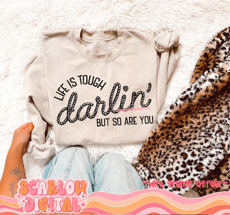 May include: A beige sweatshirt with the text "Life is tough darlin' but so are you" printed on it. The text is in a rope-like font.