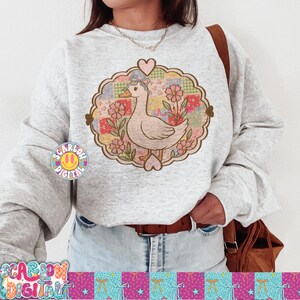 May include: A light gray sweatshirt featuring a whimsical design of a duck with a bow, surrounded by flowers and a patchwork background. The design is framed by a heart-shaped border. The sweatshirt also has the text "SCARLOW DIGITAL" and a smiley face logo.