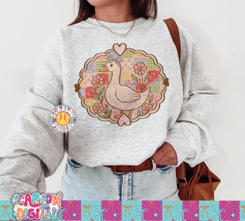 May include: A light gray sweatshirt featuring a whimsical design of a duck with a bow, surrounded by flowers and a patchwork background. The design is framed by a heart-shaped border. The sweatshirt also has the text "SCARLOW DIGITAL" and a smiley face logo.