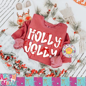 以下が含まれることがあります： 白い文字で「Holly Jolly」と書かれた赤いスウェットシャツ。赤いリボンで飾られています。緑、赤いベリー、トナカイのスリッパ、靴下などのお祝いの装飾が画像に含まれており、ホリデーテーマを作成しています。