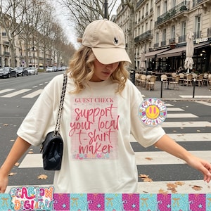 Pode incluir: Camiseta creme com o texto rosa "Guest Check Support Your Local T-Shirt Maker". Um boné de beisebol bege com um logotipo preto é usado. Uma bolsa preta com alça de corrente é visível.