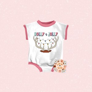 Puede incluir: Un body blanco para beb&eacute; con un ribete rosa y rojo. El body tiene un gr&aacute;fico de un reno con cuernos y luces de Navidad que dice "Holly & Jolly".