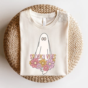 May include: Cream-colored t-shirt featuring a cartoon ghost with the words "Spooky Vibes" and a floral design in pink, yellow, and orange. The shirt is made of a soft material and is suitable for Halloween.