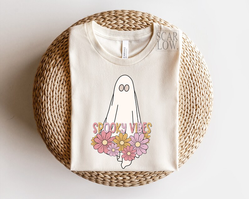 May include: Cream-colored t-shirt featuring a cartoon ghost with the words "Spooky Vibes" and a floral design in pink, yellow, and orange. The shirt is made of a soft material and is suitable for Halloween.