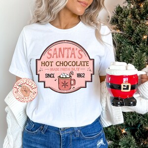 Op de afbeelding: Wit T-shirt met een roze en witte afbeelding met de tekst "Santa's Hot Chocolate Made Fresh Daily Since 1862". De afbeelding toont een mok met warme chocolademelk met marshmallows.