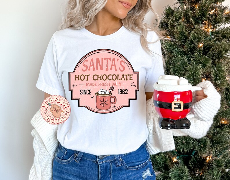 Op de afbeelding: Wit T-shirt met een roze en witte afbeelding met de tekst "Santa's Hot Chocolate Made Fresh Daily Since 1862". De afbeelding toont een mok met warme chocolademelk met marshmallows.