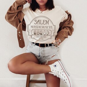 Może przedstawiać: Biały t-shirt z nadrukiem graficznym vintage'owego szyldu apteki z napisem "Salem Massachusetts Apothecary Est 1692".