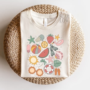 K&ouml;nnte beinhalten: Ein cremefarbenes T-Shirt mit einem Kreisdesign, das verschiedene sommerliche Illustrationen zeigt, darunter eine Wassermelone, ein Eis am Stiel, ein Strandball, Flip-Flops, ein Seestern, eine Palme, eine Muschel, ein Rettungsring und ein Flamingo.