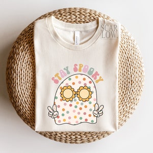 May include: A white t-shirt with a cartoon ghost wearing sunglasses and the text "Stay Spooky" in a retro font.