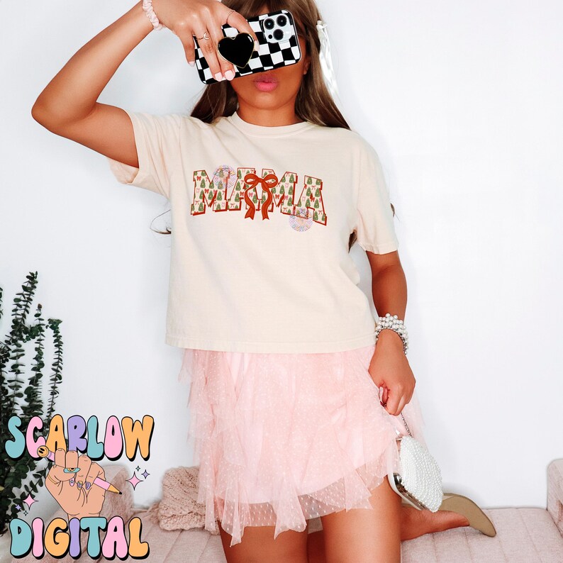 May include: A cream-colored t-shirt with the word "MAMA" printed in brown letters with a red bow and a green and brown Christmas tree pattern. The shirt is being worn by a person with a pink tulle skirt.