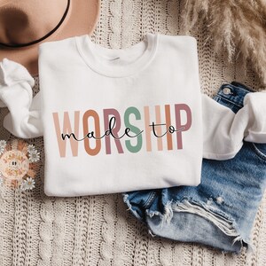 Op de afbeelding: Witte trui met de tekst "made to worship" in kleurrijke, verticale letters. De letters zijn in tinten roze, oranje, bruin, groen en perzik.