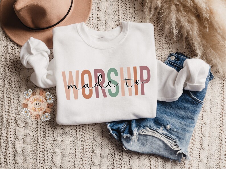 Op de afbeelding: Witte trui met de tekst "made to worship" in kleurrijke, verticale letters. De letters zijn in tinten roze, oranje, bruin, groen en perzik.