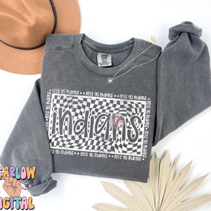 May include: A grey sweatshirt featuring the word "Indians" in a white and black checkered pattern. The phrase "lets go indians" is repeated around the design. A brown hat and gold necklace are also visible.