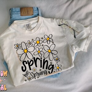 Puede incluir: Camiseta color crema con un dise&ntilde;o floral y el texto "Spring has Sprung" en escritura negra. La camiseta se combina con unos vaqueros azules y gafas de montura redonda. El dise&ntilde;o presenta margaritas negras y amarillas.