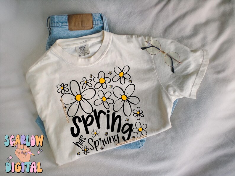 Puede incluir: Camiseta color crema con un dise&ntilde;o floral y el texto "Spring has Sprung" en escritura negra. La camiseta se combina con unos vaqueros azules y gafas de montura redonda. El dise&ntilde;o presenta margaritas negras y amarillas.