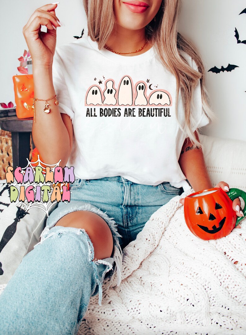 May include: A white t-shirt with a graphic of five ghosts and the text "ALL BODIES ARE BEAUTIFUL". The model is wearing the shirt with blue jeans and holding a pumpkin-shaped mug.