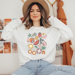 K&ouml;nnte beinhalten: Wei&szlig;er Pullover mit einem bunten, sommerlichen Grafikdruck, der eine Wassermelone, einen Strandball, einen Seestern, ein Eis am Stiel und andere sommerliche Symbole zeigt. Der Text "SCAR LOW." ist in der unteren rechten Ecke des Bildes sichtbar.