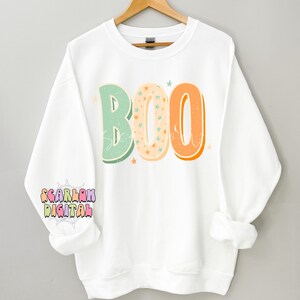 Peut inclure: Sweat-shirt blanc avec le mot "BOO" en lettres de style r&eacute;tro avec des couleurs orange, verte et cr&egrave;me. Les lettres sont remplies de petites &eacute;toiles blanches.