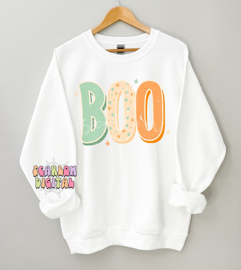 Peut inclure: Sweat-shirt blanc avec le mot "BOO" en lettres de style r&eacute;tro avec des couleurs orange, verte et cr&egrave;me. Les lettres sont remplies de petites &eacute;toiles blanches.